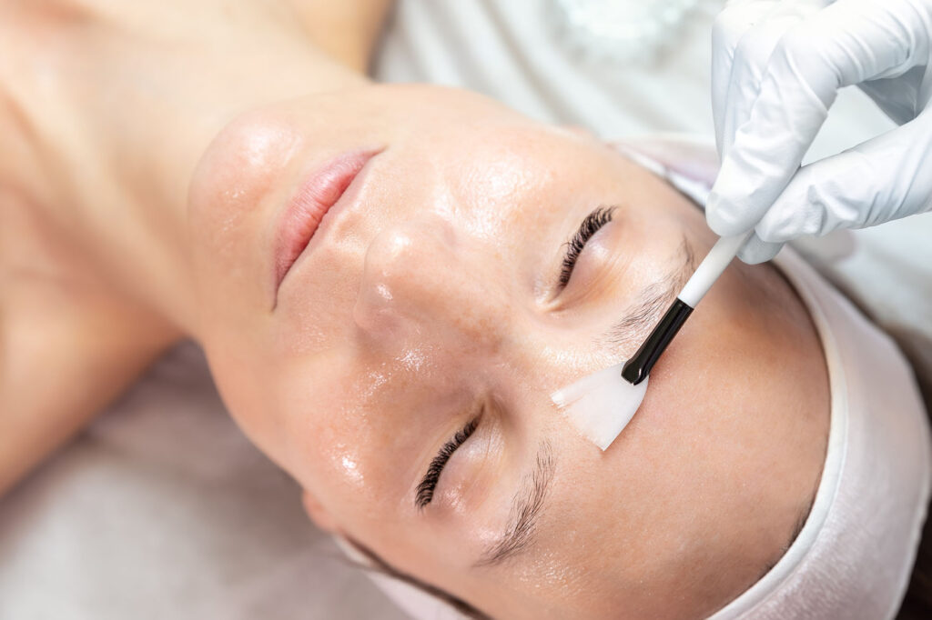 esthetician wearing gloves applying a liquid to a woman’s forehead with a small brush