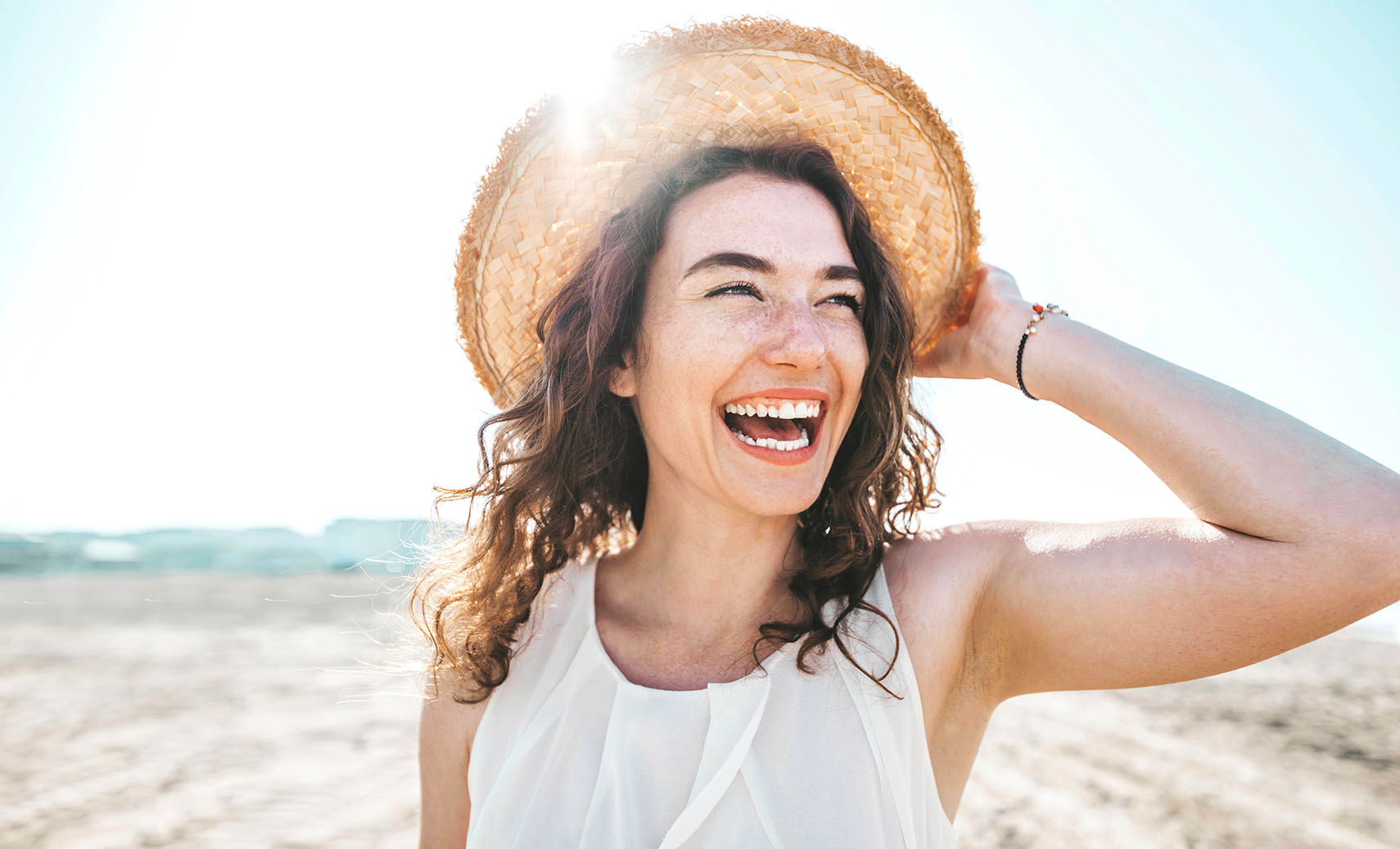 woman with curly hair wearing a straw hat and white top smiling under bright sunlight at the beach