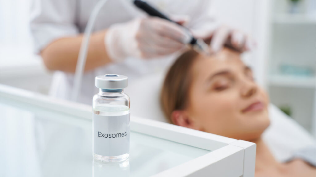 vial labeled “Exosomes” on a tray in the foreground with a practitioner performing a tool-based facial procedure on a person in the background
