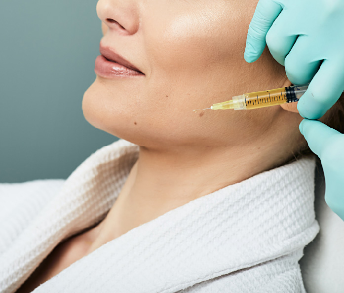close-up of a practitioner holding a syringe filled with yellow fluid near a person’s cheek while wearing teal gloves