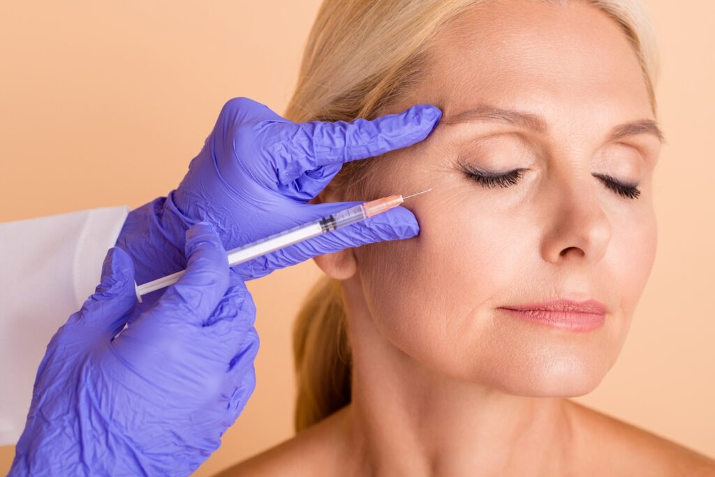 practitioner wearing purple gloves holding a syringe near a woman’s outer eye as she sits with eyes closed