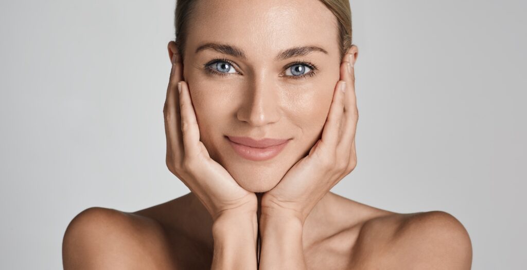 woman facing forward with both hands resting against her cheeks against a plain light background